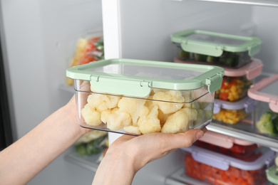 Photo of Woman taking container with frozen cauliflower cabbage from refrigerator, closeup