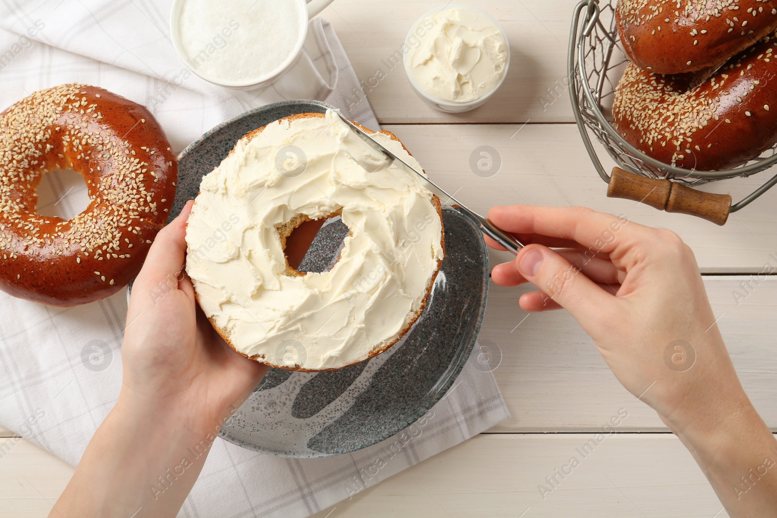 Photo of Woman making bagel with cream cheese at white wooden table, top view