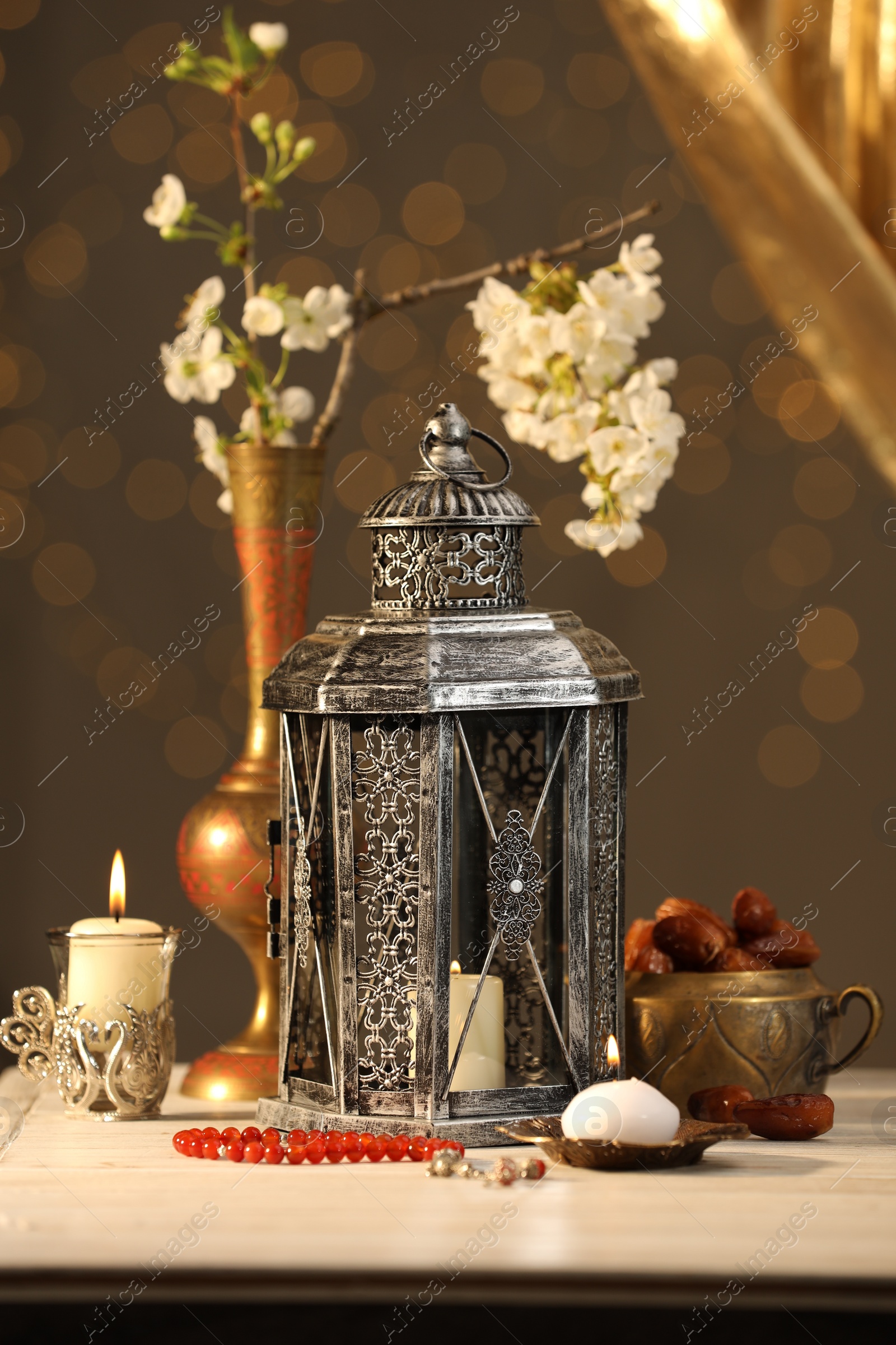 Photo of Arabic lantern, misbaha, candles, dates and flowers on table against blurred lights