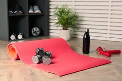 Photo of Exercise mat, dumbbells, bottle of water and skipping rope on wooden floor indoors