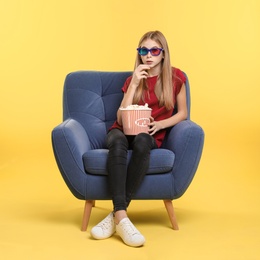 Emotional teenage girl with 3D glasses and popcorn sitting in armchair during cinema show on color background