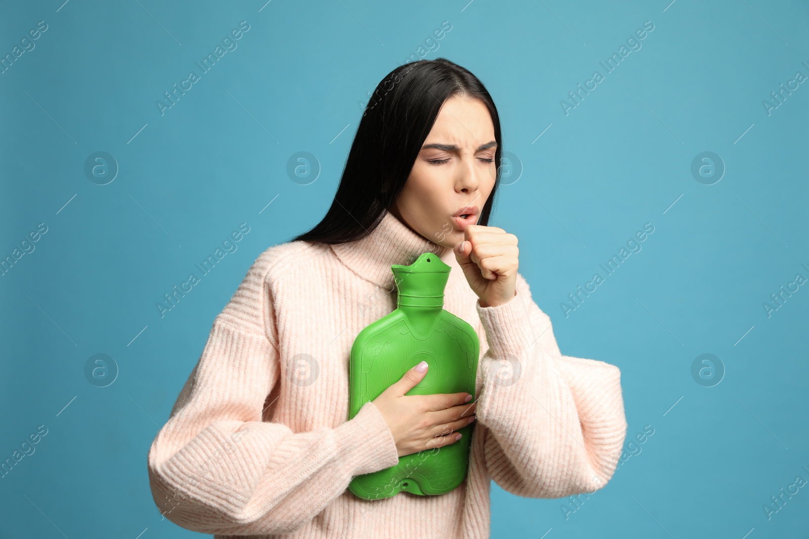 Photo of Ill woman with hot water bottle coughing on light blue background