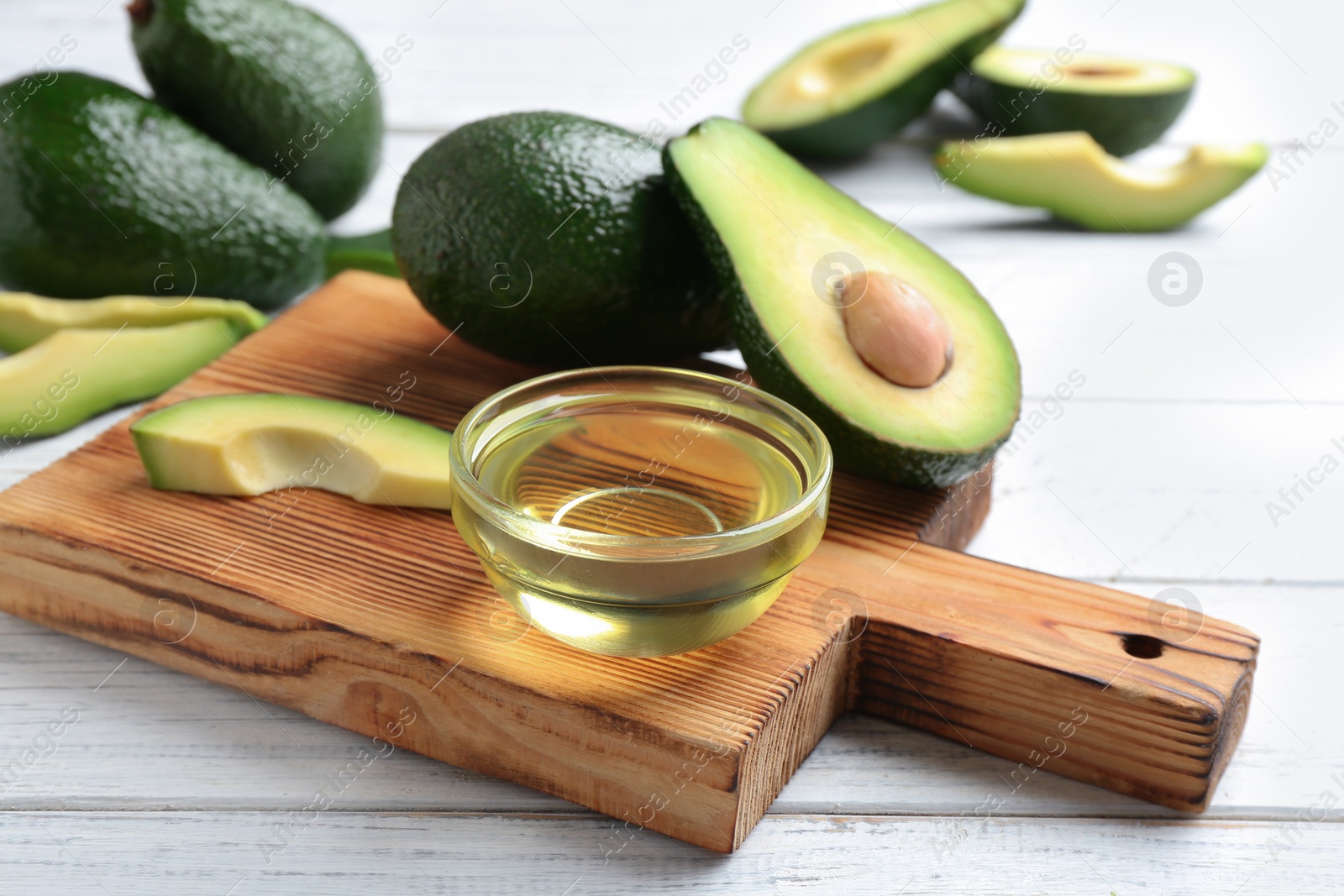 Photo of Board with bowl of natural oil and avocados on white wooden background