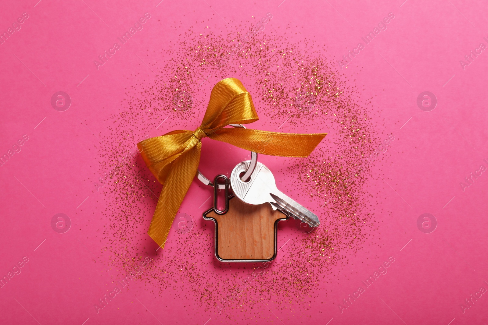 Photo of Key with trinket in shape of house, bow and glitter on pink background, flat lay. Housewarming party