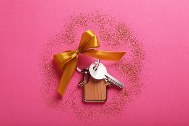 Photo of Key with trinket in shape of house, bow and glitter on pink background, flat lay. Housewarming party