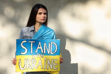 Photo of Sad woman holding poster in colors of national flag and words Stand with Ukraine near light wall. Space for text