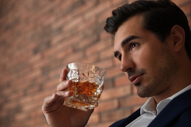 Young man with glass of whiskey near brick wall indoors. Space for text