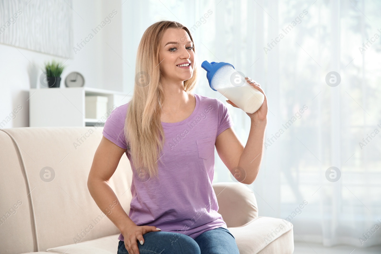 Photo of Young woman with bottle of protein shake sitting on sofa at home