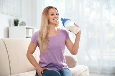 Young woman with bottle of protein shake sitting on sofa at home