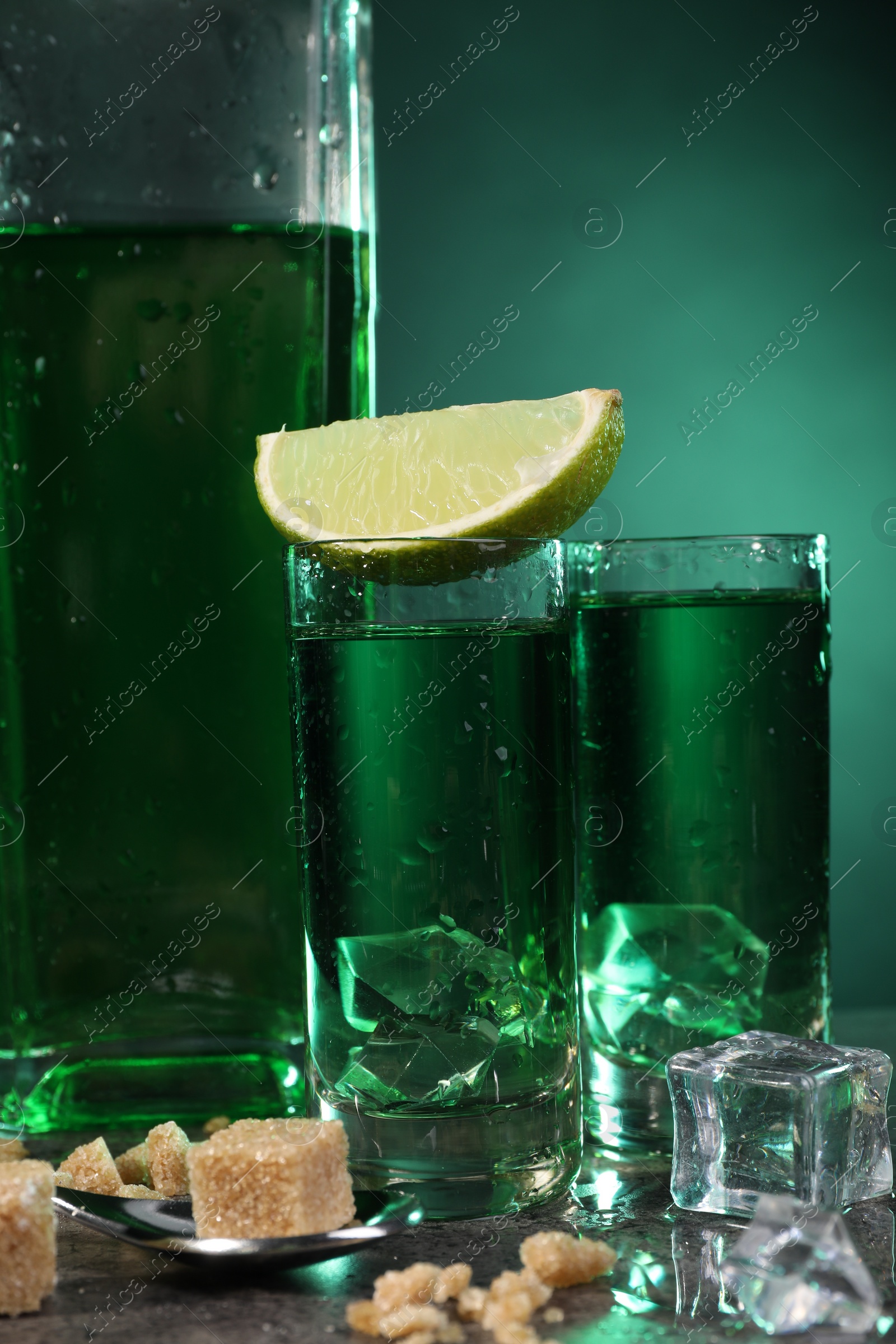 Photo of Absinthe in shot glasses with ice cubes, lime, brown sugar and spoon on gray table, closeup. Alcoholic drink