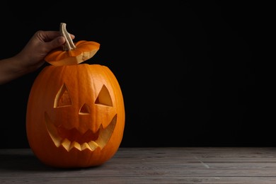 Woman with scary jack o'lantern made of pumpkin on wooden table against black background, space for text. Halloween traditional decor