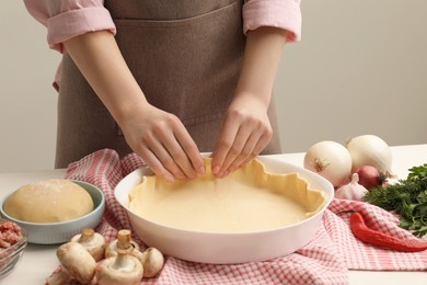 Photo of Woman putting dough for meat pie into baking dish at white table, closeup