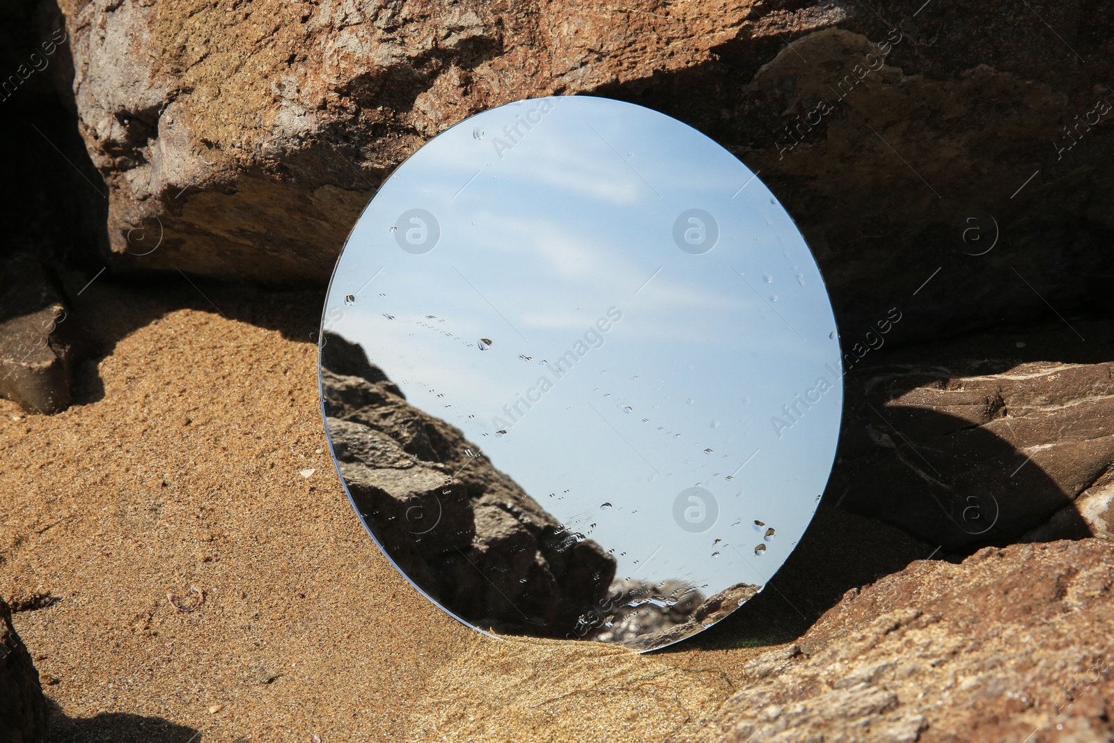 Photo of Round mirror reflecting light blue sky on sand near stones outdoors