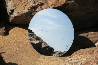 Round mirror reflecting light blue sky on sand near stones outdoors