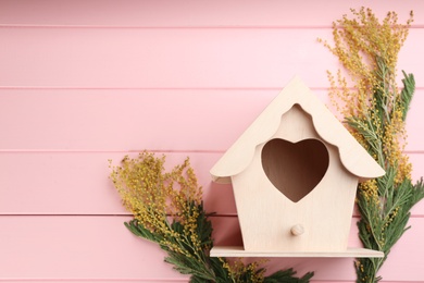 Beautiful bird house with heart shaped hole and mimosa flowers on pink wooden background, space for text. Spring flat lay composition