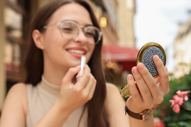 Photo of Beautiful young woman with cosmetic pocket mirror applying lipstick outdoors, focus on hand