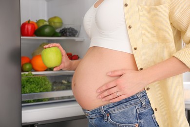 Photo of Young pregnant woman with apple near fridge at home, closeup. Healthy eating