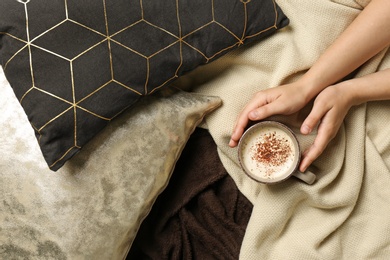 Photo of Woman holding cup of coffee while lying on bed with pillows and warm plaid, top view