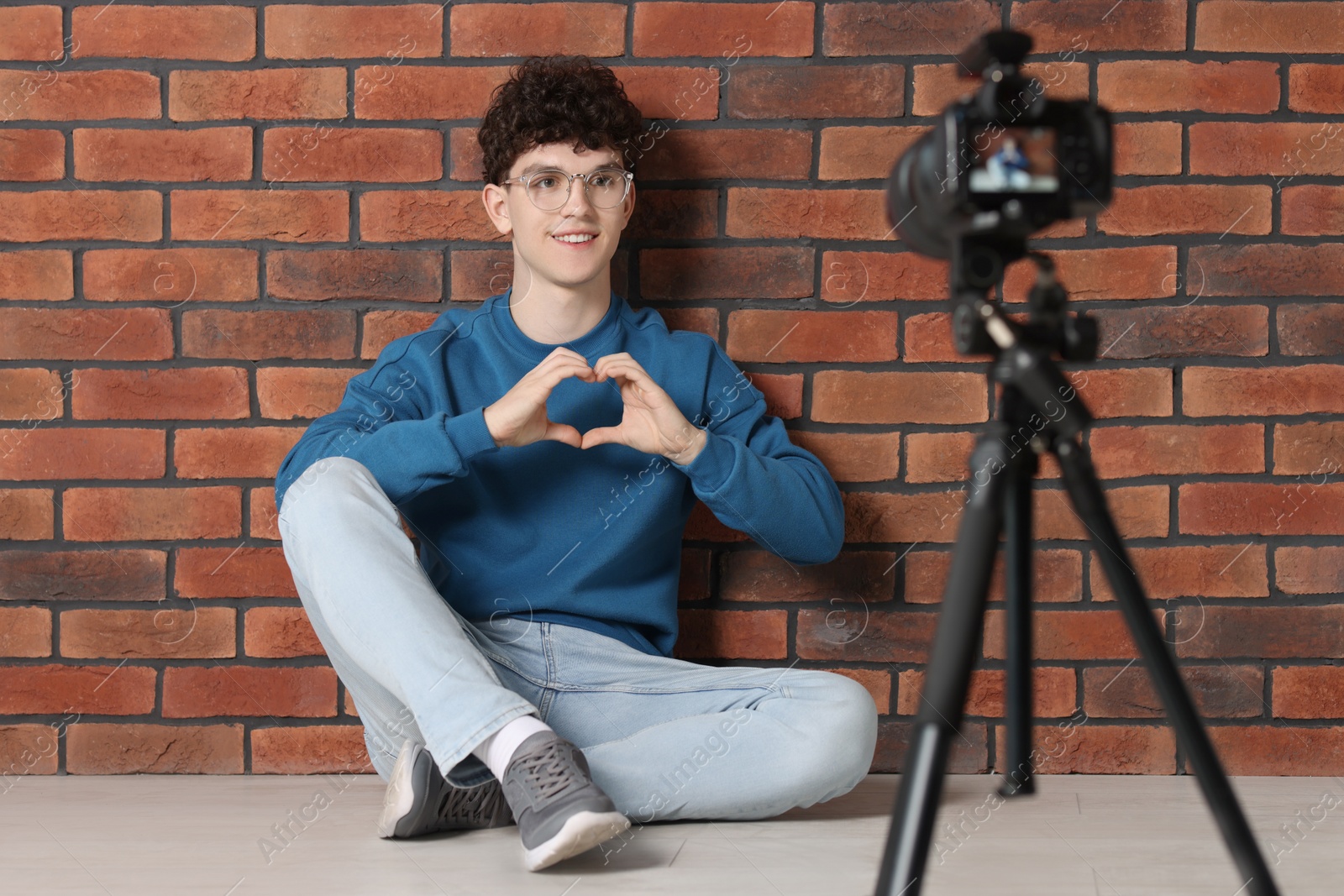 Photo of Smiling teenage blogger making heart gesture to his subscribers while streaming indoors