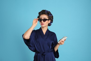 Young woman in silk bathrobe with hair curlers, sunglasses and smartphone on light blue background