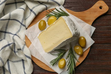 Photo of Board with piece of tasty camembert cheese, olives and rosemary on wooden table, top view