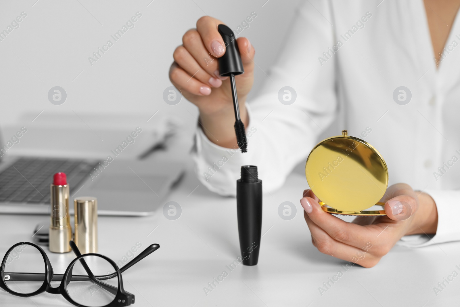 Photo of Young woman using cosmetic pocket mirror and mascara at white table indoors, closeup