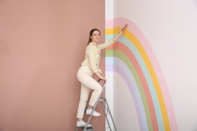 Photo of Young woman painting rainbow on white wall indoors