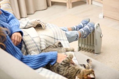 Photo of Young woman with cat warming feet on electric heater at home, closeup