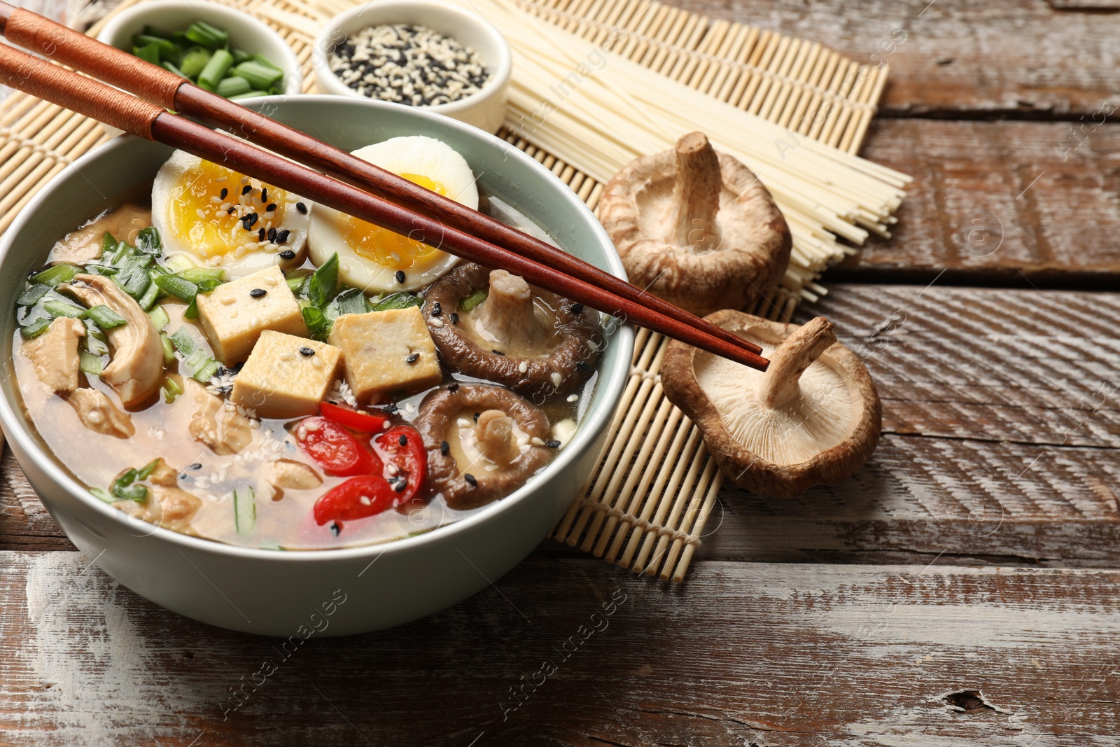 Photo of Noodle soup. Bowl of delicious ramen, ingredients and chopsticks on wooden table, closeup. Space for text