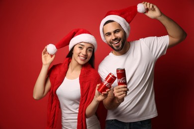 Photo of MYKOLAIV, UKRAINE - JANUARY 27, 2021: Young couple in Christmas hats holding cans of Coca-Cola on red background