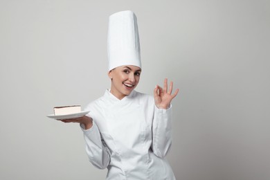 Happy professional confectioner in uniform holding delicious cheesecake on light grey background