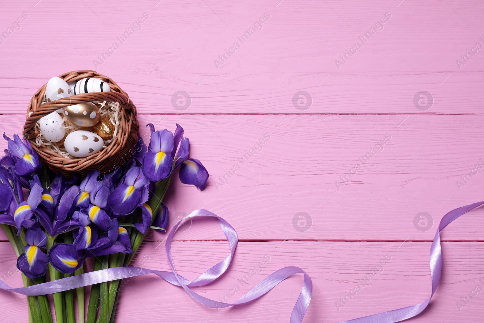 Photo of Flat lay composition with festively decorated Easter eggs in wicker basket and iris flowers on pink wooden background. Space for text