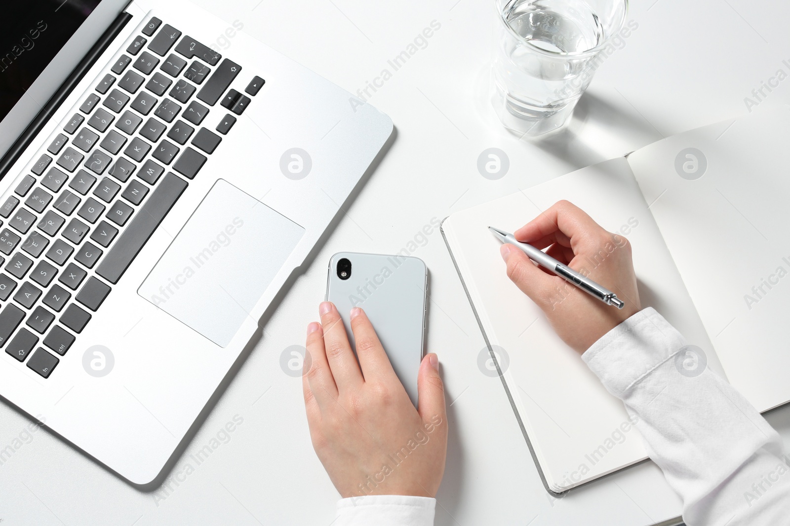 Photo of Woman with smartphone writing in notebook at white table, closeup