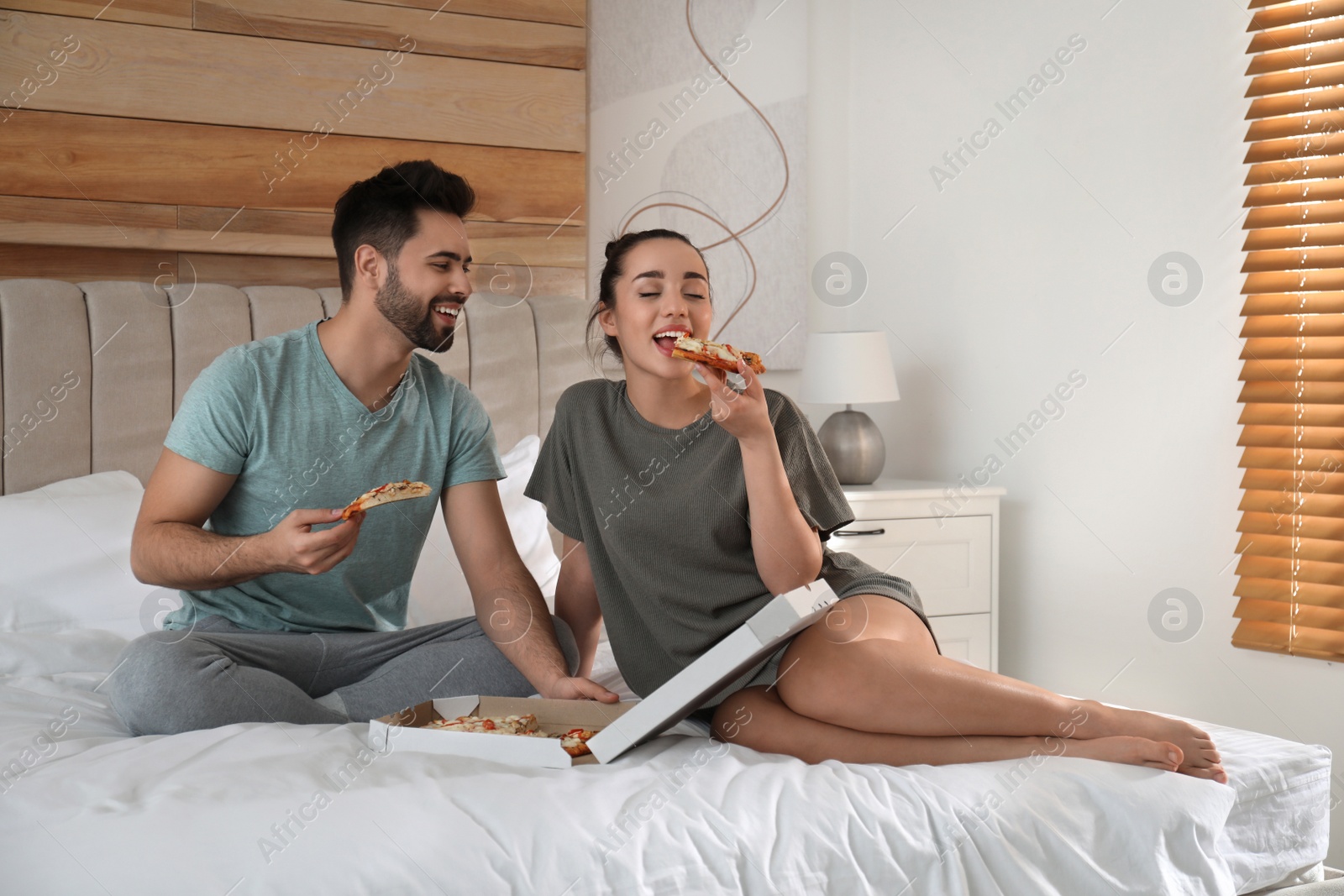 Photo of Happy couple in pajamas eating pizza on bed at home