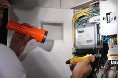 Electrician with flashlight fixing electric panel indoors, closeup