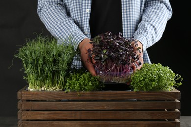 Man with wooden crate of different fresh microgreens on black background, closeup