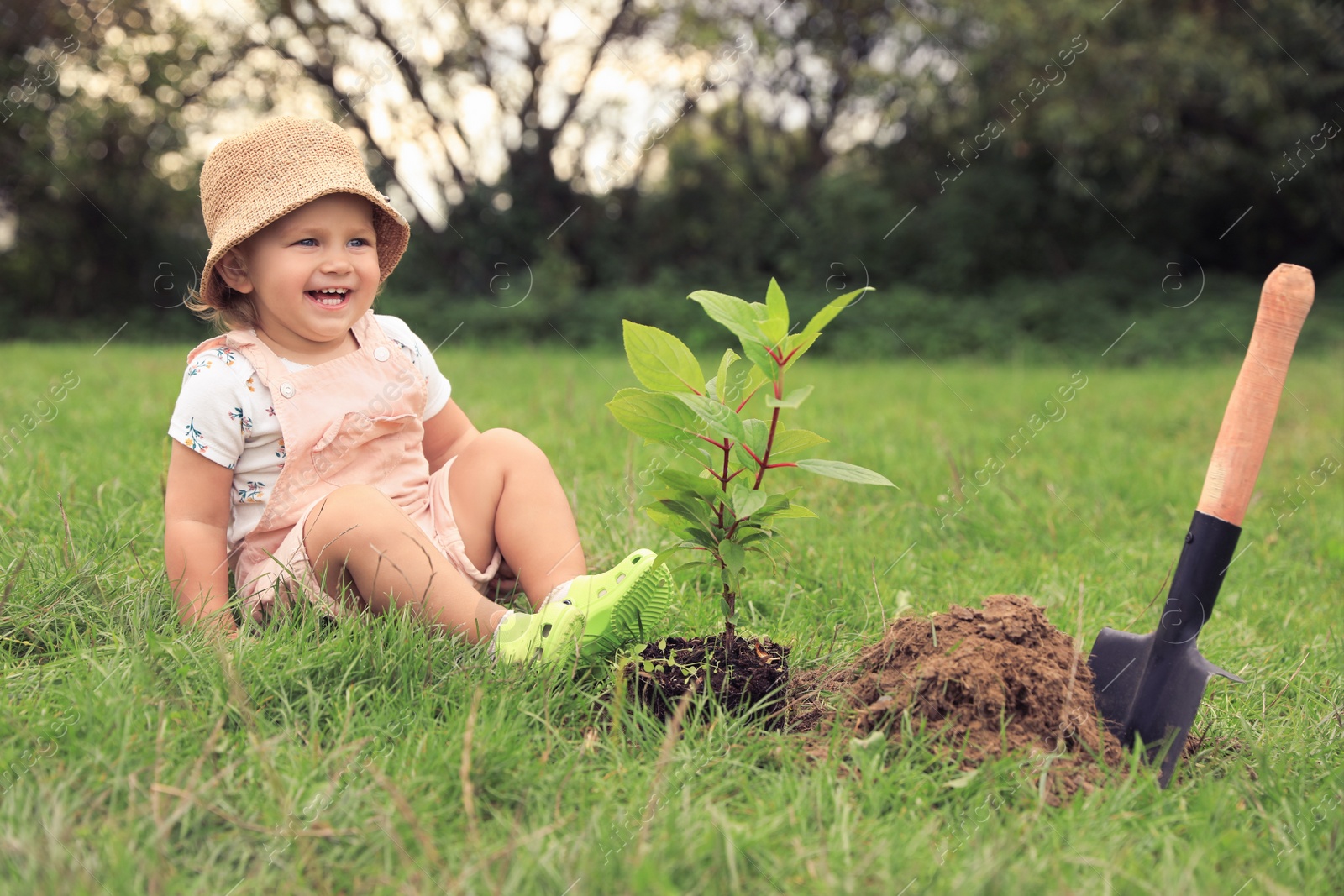 Photo of Cute happy baby girl near young green tree and trowel in garden