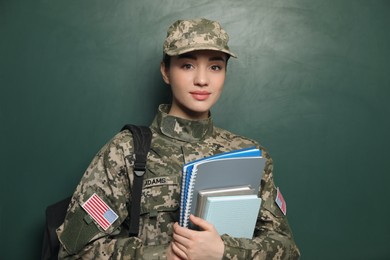 Photo of Female soldier with notebooks and backpack near green chalkboard. Military education