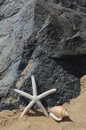 Beautiful starfish and sea shell near black stone on sandy beach, space for text