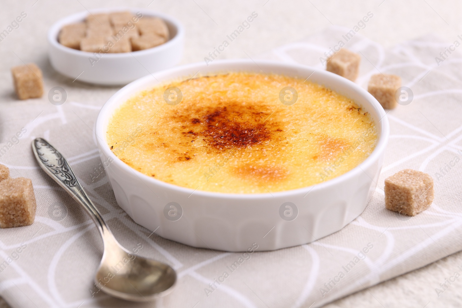 Photo of Delicious creme brulee in bowl, sugar cubes and spoon on table, closeup