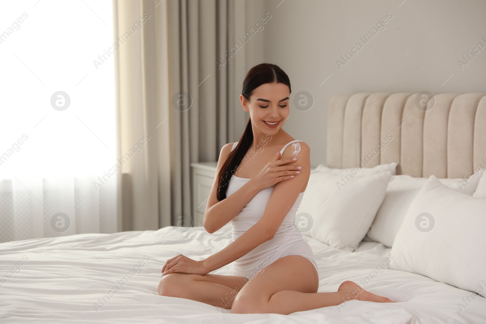 Photo of Young woman applying body cream onto her shoulder at home