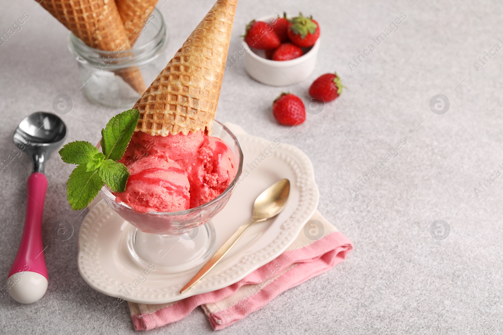 Photo of Delicious scoops of strawberry ice cream with mint and wafer cone in glass dessert bowl served on grey table. Space for text