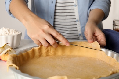 Photo of Woman cutting dough leftovers for traditional English apple pie in baking dish at table, closeup