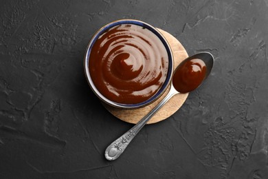 Photo of Tasty barbeque sauce in bowl and spoon on dark textured table, top view