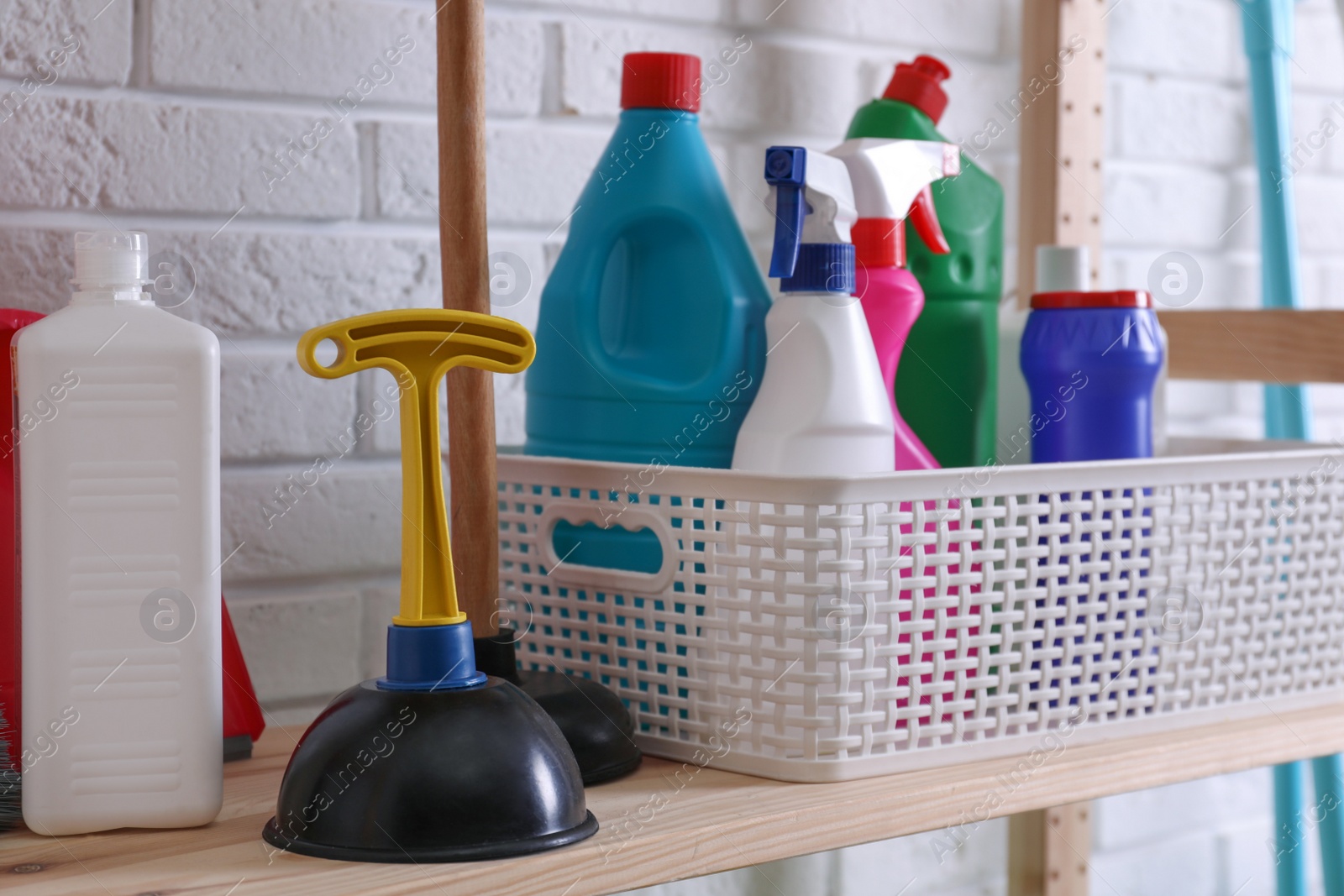 Photo of Plungers and basket with detergents on wooden shelf near white brick wall