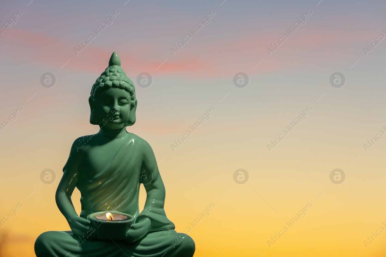 Photo of Decorative Buddha statue with burning candle outdoors at sunset. Space for text