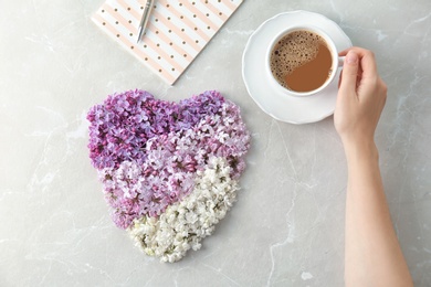 Woman with cup of coffee near heart made of blossoming lilac on light background, top view. Spring flowers