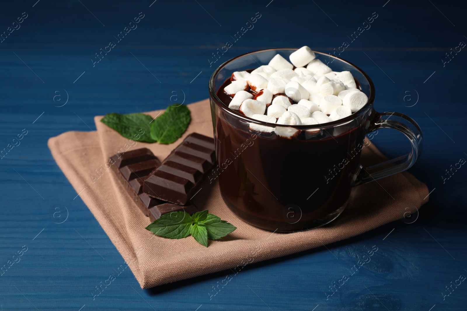 Photo of Glass cup of delicious hot chocolate with marshmallows and fresh mint on blue wooden table