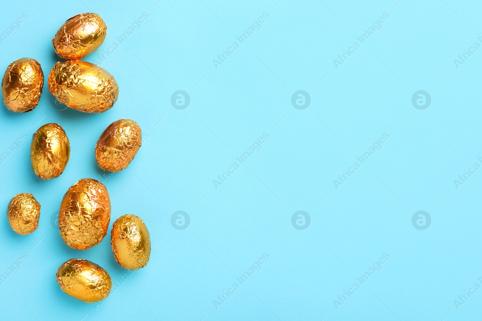 Photo of Chocolate eggs wrapped in golden foil on light blue background, flat lay. Space for text