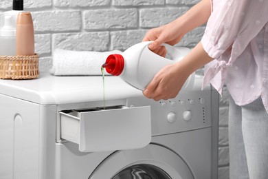 Photo of Woman pouring fabric softener from bottle into washing machine near white brick wall, closeup